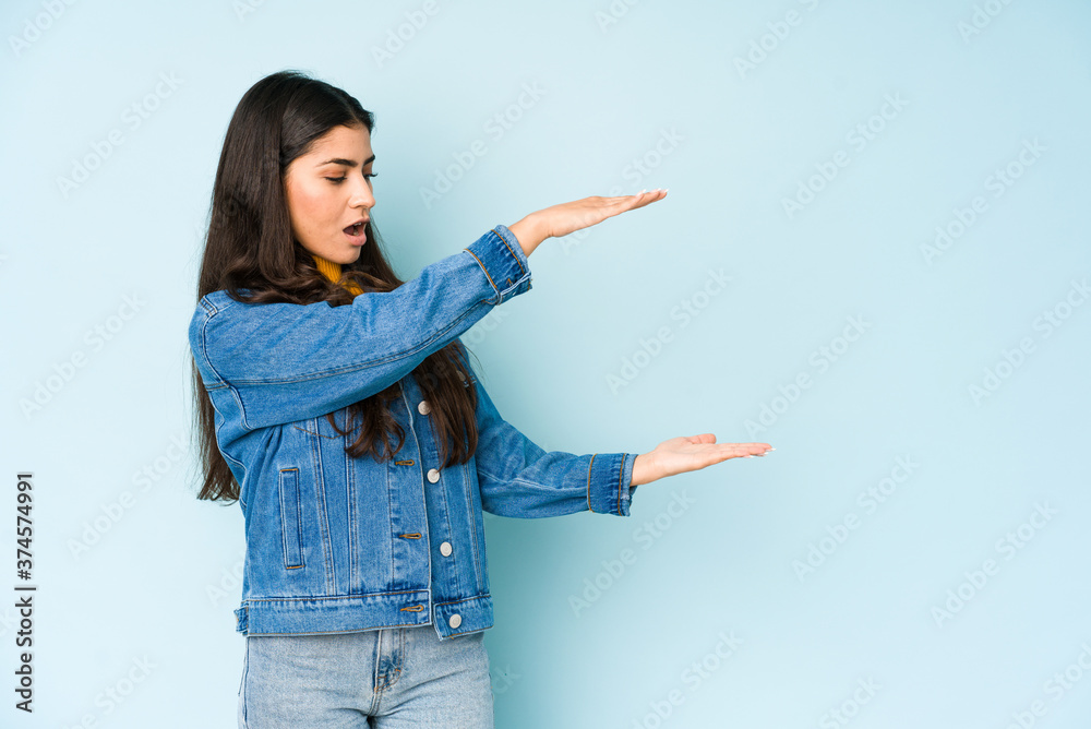 Young indian woman isolated on blue background shocked and amazed holding a copy space between hands. Young indian woman isolated on blue background shocked and amazed holding a copy space between hands.