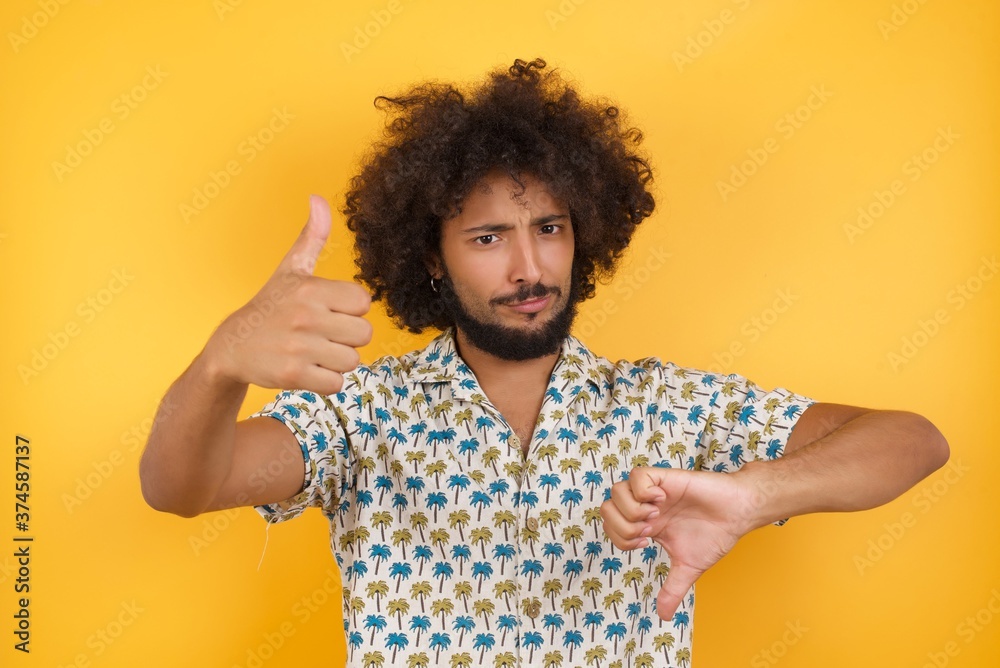 Young man with afro hair over wearing hawaiian shirt standing over yellow background showing thumbs up and thumbs down, difficult choose concept Young man with afro hair over wearing hawaiian shirt standing over yellow background showing thumbs up and thumbs down, difficult choose concept