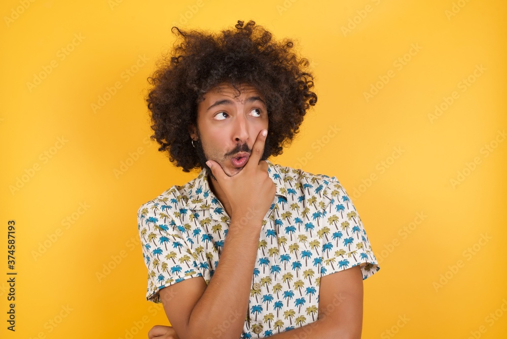 Young man with afro hair over wearing hawaiian shirt standing over yellow background Thinking worried about a question, concerned and nervous with hand on chin. Young man with afro hair over wearing hawaiian shirt standing over yellow background Thinking worried about a question, concerned and nervous with hand on chin.