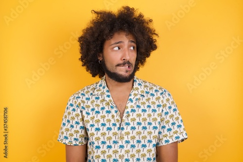 Photo of amazed Young man with afro hair over wearing hawaiian shirt standing over yellow background bitting lip and looking up to empty space,