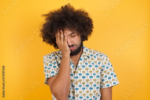 Tired overworked Young man with afro hair over wearing hawaiian shirt standing over has sleepy expression, gloomy look, covers face with hand, has eyes shut, gasps from tiredness, fatigue after party