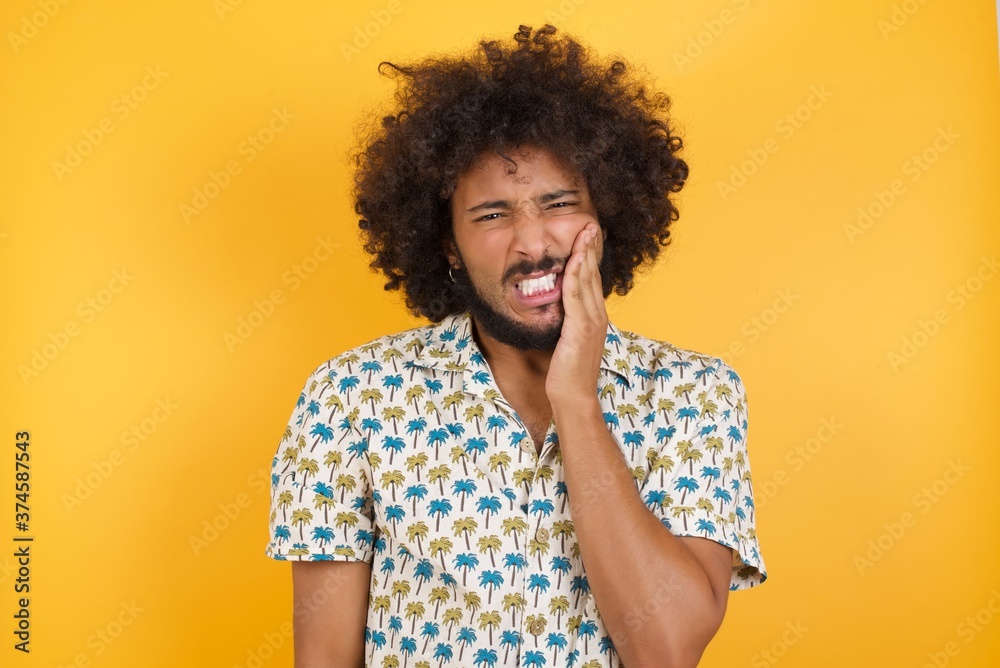 Young man with afro hair over wearing hawaiian shirt standing over yellow background touching mouth with hand with painful expression because of toothache or dental illness on teeth. Young man with afro hair over wearing hawaiian shirt standing over yellow background touching mouth with hand with painful expression because of toothache or dental illness on teeth.