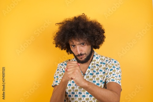 Young man with afro hair over wearing hawaiian shirt standing over yellow background Ready to fight with fist defense gesture, angry and upset face, afraid of problem. © Roquillo