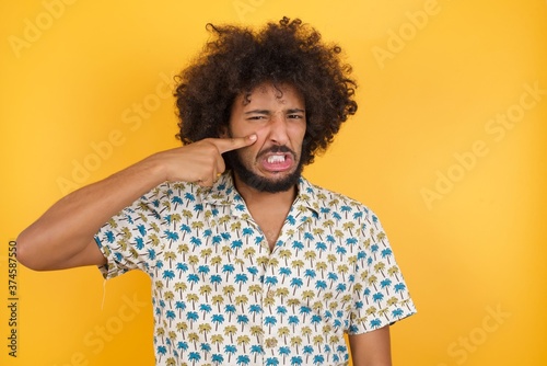 Young man with afro hair over wearing hawaiian shirt standing over yellow background pointing unhappy to pimple on forehead, ugly infection of blackhead. Acne and skin problem