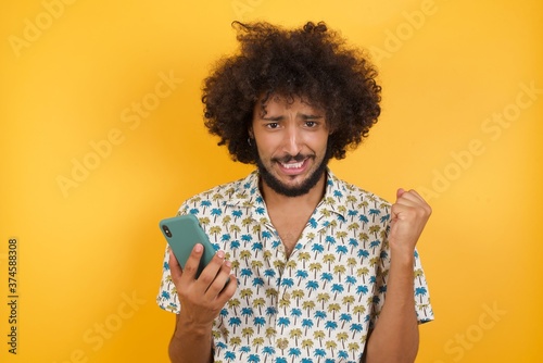 Positive hipster Young man with afro hair over wearing hawaiian shirt standing over yellow wall holds modern cell phone connected to headphones, clenches fist from good emotions, exclaims with joy,