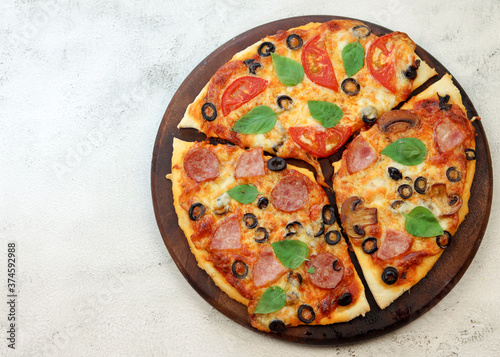 Pizza with sausage, ham, olives and mushrooms on a round wooden cutting board on a light gray background. Top view, flat lay