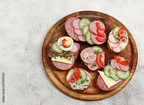 Assorted sandwiches with vegetables and sausages on the round wooden cutting board on a light gray background. Top view, flat lay
