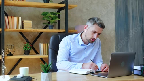 Gray-haired bearded focused man at home office in wireless earphones looks laptop computer writes information notebook. Guy study remotely online video call conference with teacher, distance education photo