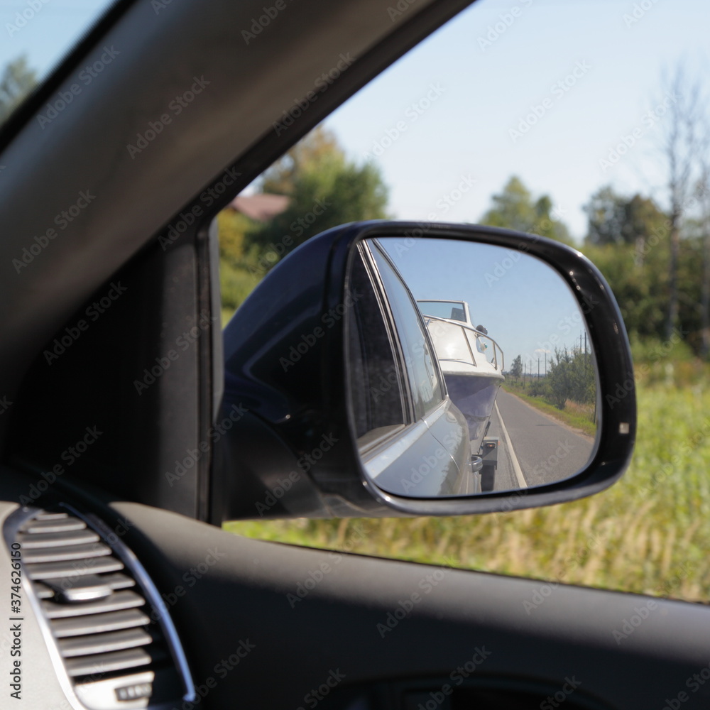 Car rear wiew side mirror with boat on trailer reflection and field and trees background, boats transportation, outdoor active recreation tourism on Sunny summer day Car rear wiew side mirror with boat on trailer reflection and field and trees background, boats transportation, outdoor active recreation tourism on Sunny summer day