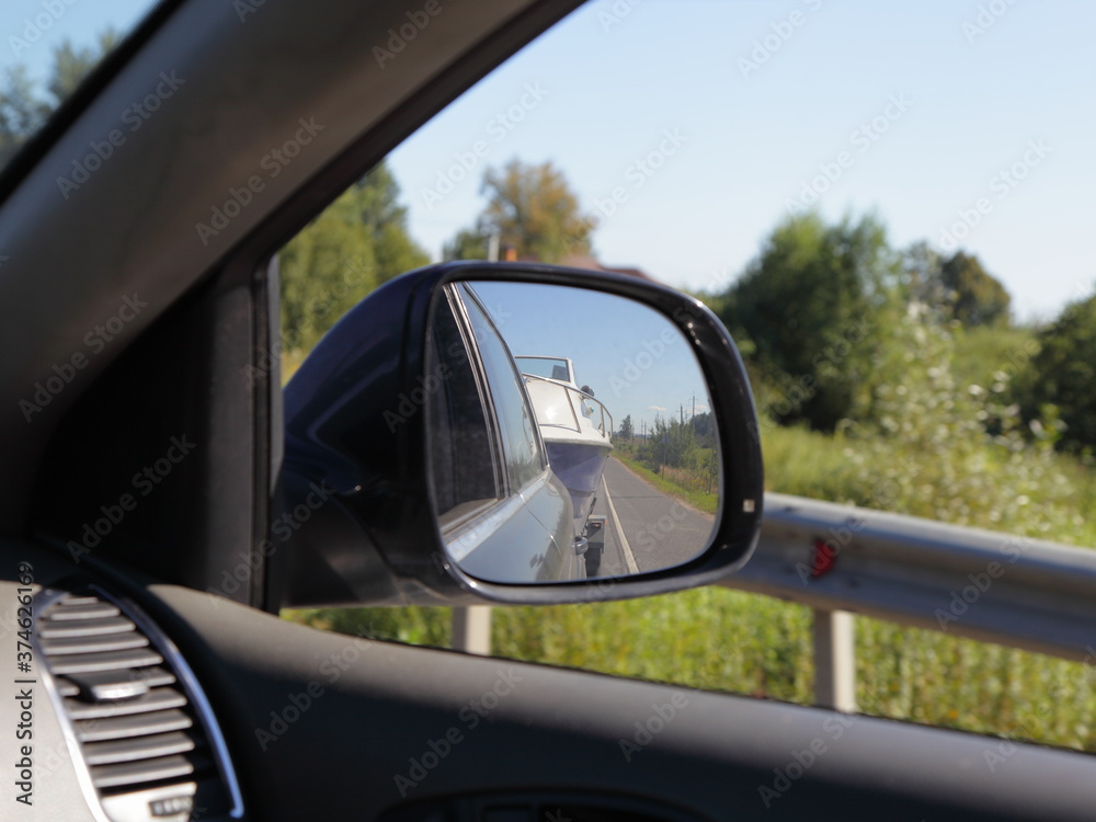 Car caravaning travel trip active vacation on Sunny summer day, indoor view from car to front side window to outdoor back view mirror reflection with boat on trailer, road fence and natural landscape