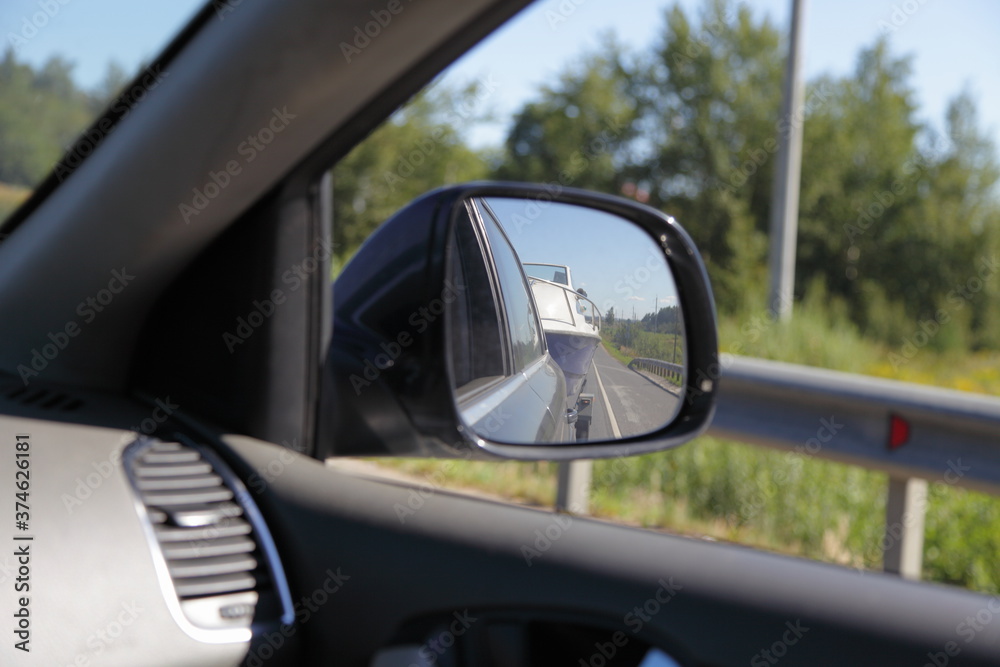 Indoor view from car in front side window to outdoor back view mirror reflection with boat on trailer, road fence and natural landscape ,automobile trip active vacation in Russia on Sunny summer day