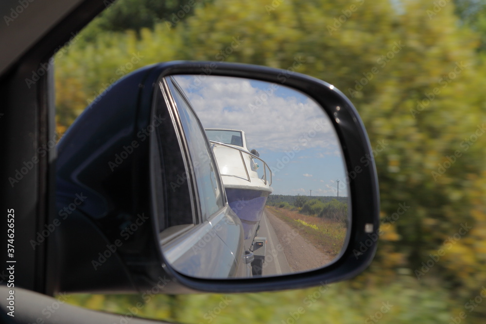 Cabin motor boat towing on car trailer, close up view in reflection in right side car rear mirrow on blue sky and green field background, travel with private boat