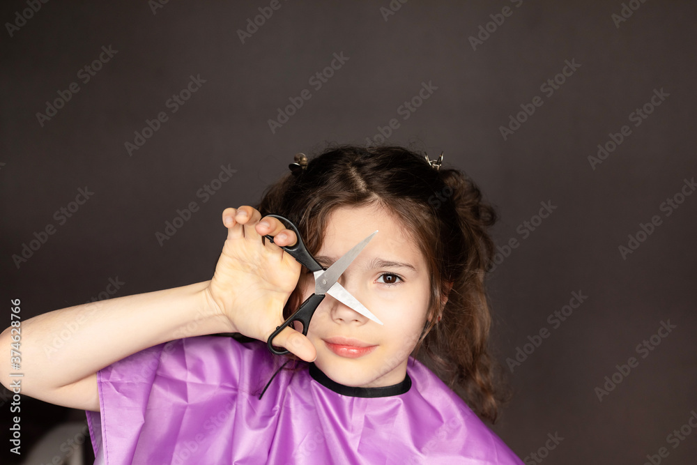 Portrait of a Caucasian young curly-haired girl holds metal scissors at her face on a dark background. The concept of a self-employed hairdresser, home Barber. DIY hair cut during covid-19 Portrait of a Caucasian young curly-haired girl holds metal scissors at her face on a dark background. The concept of a self-employed hairdresser, home Barber. DIY hair cut during covid-19