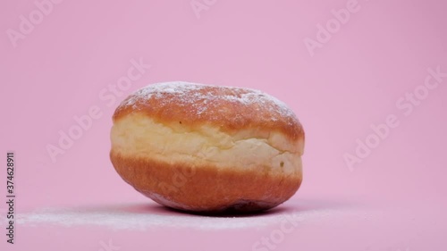 Time-lapse shot of a round donut. Donut with filling. Powdered sugar on top. Pink background.