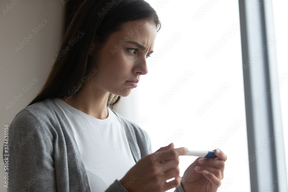 Close up anxious unhappy woman holding plastic pregnancy test kit in hands, stressed frustrated young female dissatisfied by result, health problem, infertility, unwanted pregnancy, standing at home Close up anxious unhappy woman holding plastic pregnancy test kit in hands, stressed frustrated young female dissatisfied by result, health problem, infertility, unwanted pregnancy, standing at home