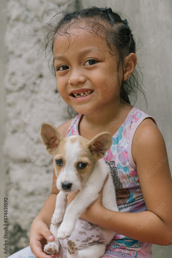 Pretty and Cute Little Girl Playing and Holding her Small Fluffy Dog Puppy at Home in Asia in the Philippines. Toothless Happy Kid with missing Teeth Pretty and Cute Little Girl Playing and Holding her Small Fluffy Dog Puppy at Home in Asia in the Philippines. Toothless Happy Kid with missing Teeth