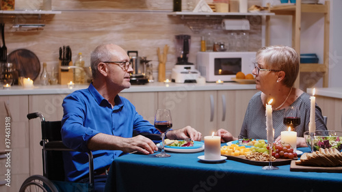 Aged senior man with disabilities having romantic dinner sitting at the table in cozy kitchen. Wheelchair immobilized paralyzed handicapped man dining with wife at home, enjoying the meal
