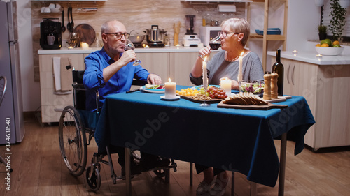 Paralyzed man toasting with wine with his wife. Senior old man in wheelchair dining with aged woman sitting in the kitchen. Imobilized paralyzed handicapped elderly husband having romantic dinner
