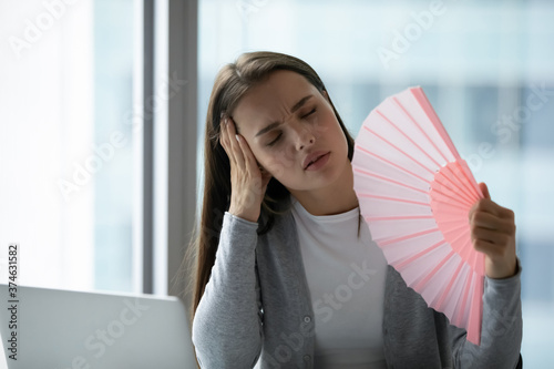 Φωτογραφία Overheated unhappy young businesswoman waving paper fan in office, sitting at de