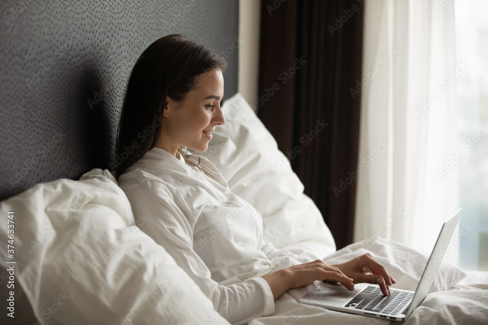 Smiling young woman wearing bathrobe using laptop, sitting in bed under warm duvet, beautiful female working online at home or in hotel, writing email, chatting in social network, relaxing Smiling young woman wearing bathrobe using laptop, sitting in bed under warm duvet, beautiful female working online at home or in hotel, writing email, chatting in social network, relaxing