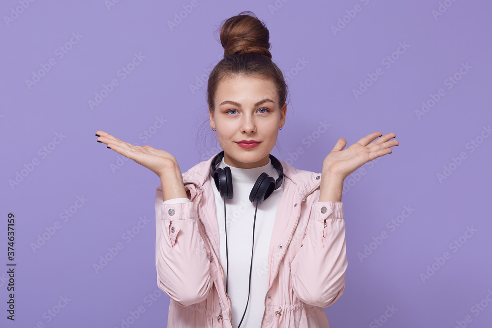 Young woman girl in rosy jacket posing isolated over lilac background, listen music, posing with headphones around neck, spreading hands aside, looks at camera. Young woman girl in rosy jacket posing isolated over lilac background, listen music, posing with headphones around neck, spreading hands aside, looks at camera.