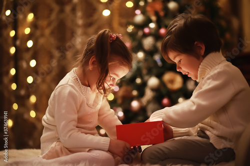 Family on Christmas eve at fireplace. Kids opening Xmas presents. Children under Christmas tree with gift boxes. Decorated living room with traditional fire place. Cozy warm winter evening at home.