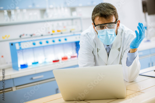 Fototapeta Naklejka Na Ścianę i Meble -  Researcher in white lab coat and protective mask working using laptop while sitting in the lab