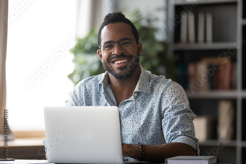 Head shot portrait smiling African American businessman wearing glasses sitting at work desk with laptop, excited freelancer looking at camera, happy student working on online research project