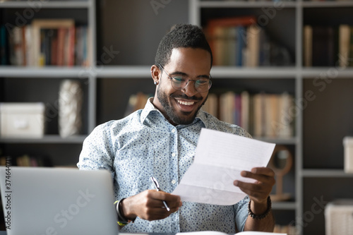 Happy smiling African American man wearing glasses reading letter at workplace, sitting at work desk, satisfied businessman received good news, job promotion, money refund or great exam results photo