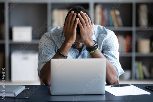 Unhappy hopeless African American man holding head in hands, overwhelmed tired businessman sitting at work desk with laptop, feeling exhausted, financial problem, loss money or bankruptcy photo