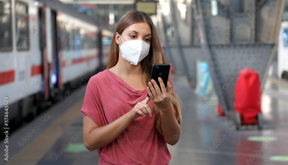 Young woman wearing KN95 FFP2 face mask waiting train at the station. Casual woman using smart phone in train station. Young woman wearing KN95 FFP2 face mask waiting train at the station. Casual woman using smart phone in train station.