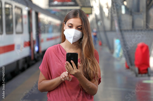 Young woman wearing KN95 FFP2 face mask waiting train at the station. Caucasian woman using smart phone in train station.