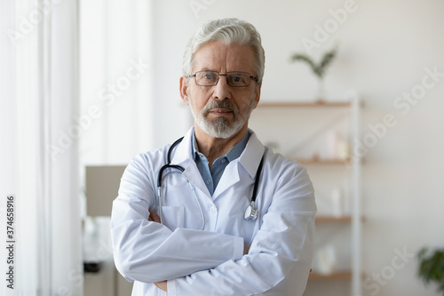 Head shot portrait mature doctor wearing glasses standing with arms crossed in office, confident serious senior therapist practitioner gp wearing white uniform coat with stethoscope looking at camera