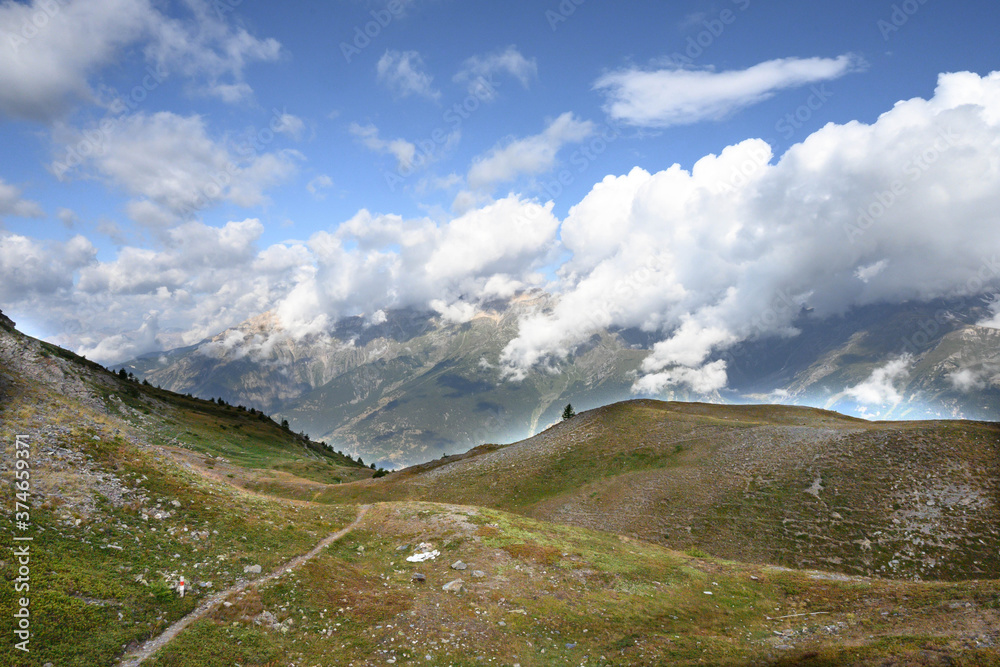 landscape of the alpine meadows, road, path, footpath, and on the horizon mountains covered by clouds and a blue sky,