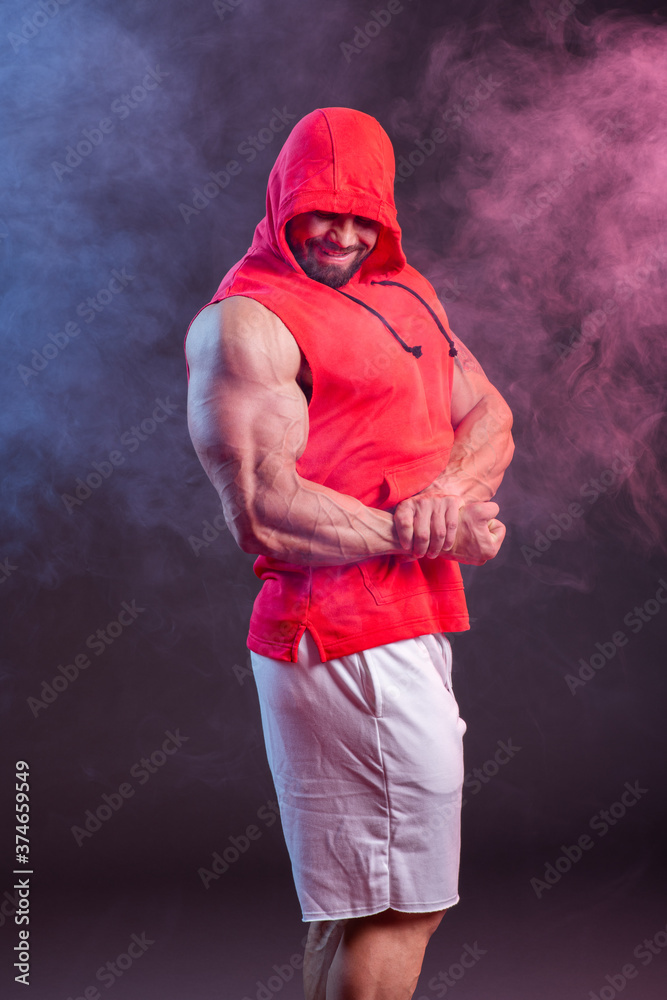 Powerful muscular man with a hood posing in the red sweatshirt. Studio shoot with red and blue smoke Powerful muscular man with a hood posing in the red sweatshirt. Studio shoot with red and blue smoke