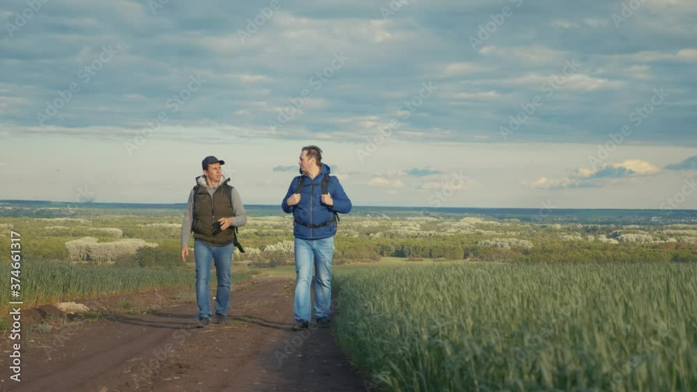 teamwork travel. two man hikers with backpacks walk lifestyle along trail next to a field with green grass in nature. concept travel adventure hiking healthy. two tourists walk walking lifestyle teamwork travel. two man hikers with backpacks walk lifestyle along trail next to a field with green grass in nature. concept travel adventure hiking healthy. two tourists walk walking lifestyle