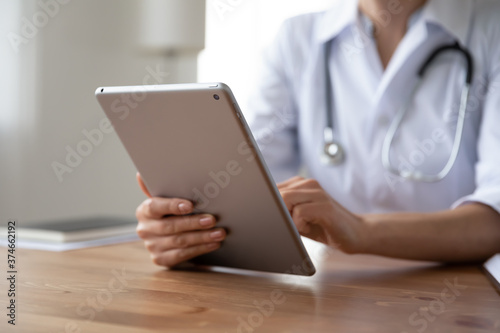 Close up young woman doctor holding digital tablet, working online, sitting at desk, female physician wearing white coat with stethoscope using medical apps, consulting patient, telemedicine