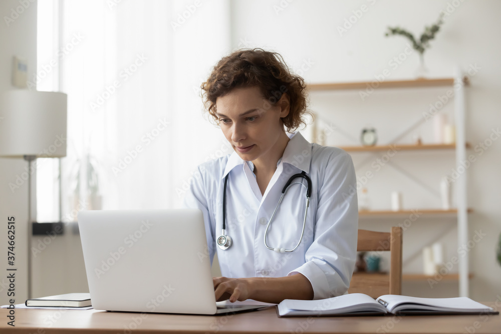 Serious woman wearing white uniform with stethoscope working on laptop, sitting at desk in hospital, focused doctor physician gp looking at computer screen, using medical apps, consulting online Serious woman wearing white uniform with stethoscope working on laptop, sitting at desk in hospital, focused doctor physician gp looking at computer screen, using medical apps, consulting online