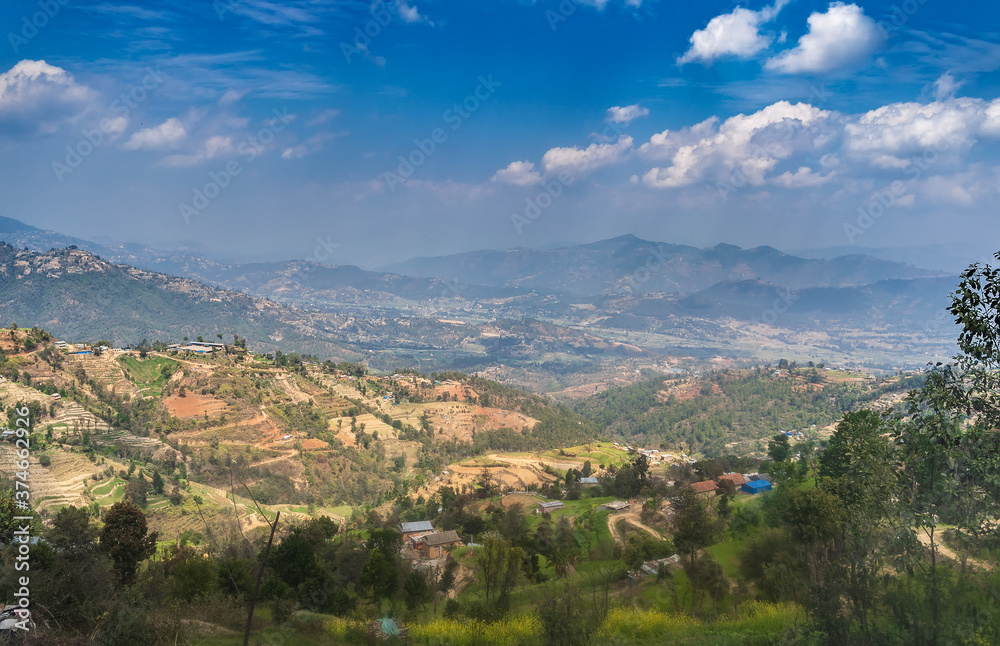 The eastern part of the Kathmandu valley, on a sunny day, with the hills and foothills of the Himalayas, terraces with agricultural crops, against the backdrop of a blue sky and clouds.