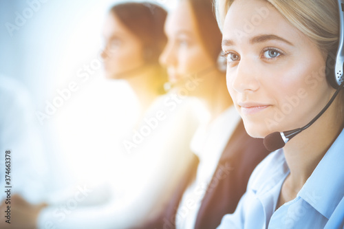 Cheerful smiling business woman with headphones consulting clients. Group of diverse phone operators at work in sunny office.Call center and business people concept
