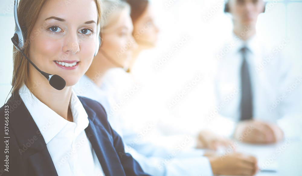Cheerful smiling business woman with headphones consulting clients. Group of diverse phone operators at work in sunny office.Call center and business people concept Cheerful smiling business woman with headphones consulting clients. Group of diverse phone operators at work in sunny office.Call center and business people concept