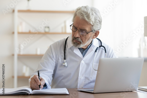 Serious mature doctor wearing glasses working with medical documents and laptop, sitting at desk in hospital, busy senior therapist practitioner gp writing illness history, filling patient card