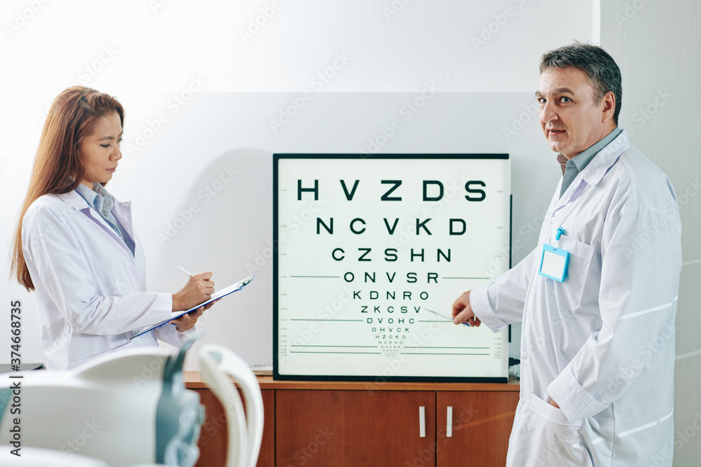 Ophthalmologist asking patient to name letters on test chart, when nurse taking notes in medical card