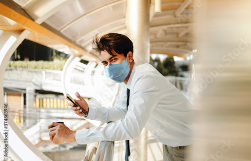 Young Asian Businessman Wearing a Surgical Mask and Using a Smart Phone in City. Healthcare in New Normal Lifestyle Concept. Looking at the Camera