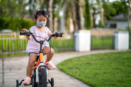 Cute little girl with protective face mask riding bicycle at village. 