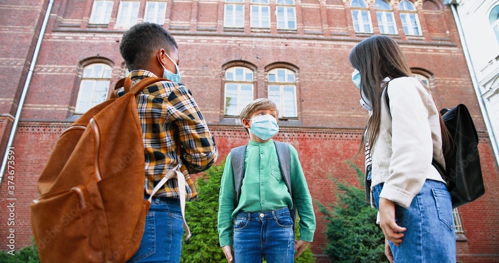 Portrait of multi-ethnic children in medical masks greeting with elbows near school in quarantine. Cute African American and Caucasian boys outdoors. Beautiful girl with friends. School concept