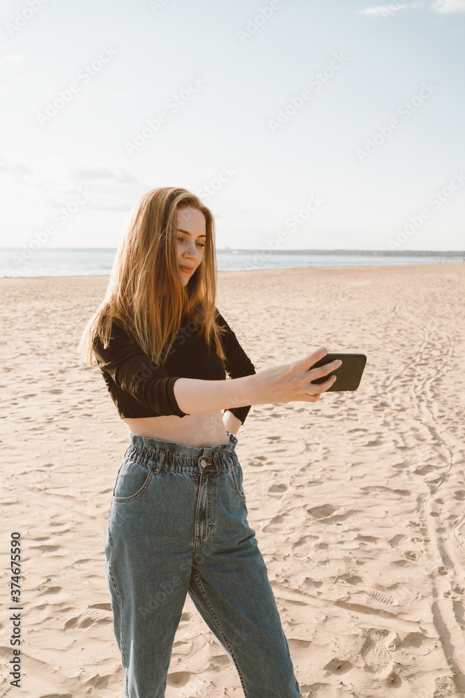 Beautiful woman taking selfie on ocean or sea coastline in sunny day. Pretty female with long hair, blonde takes photo on mobile phone on sandy beach in summer or autumn