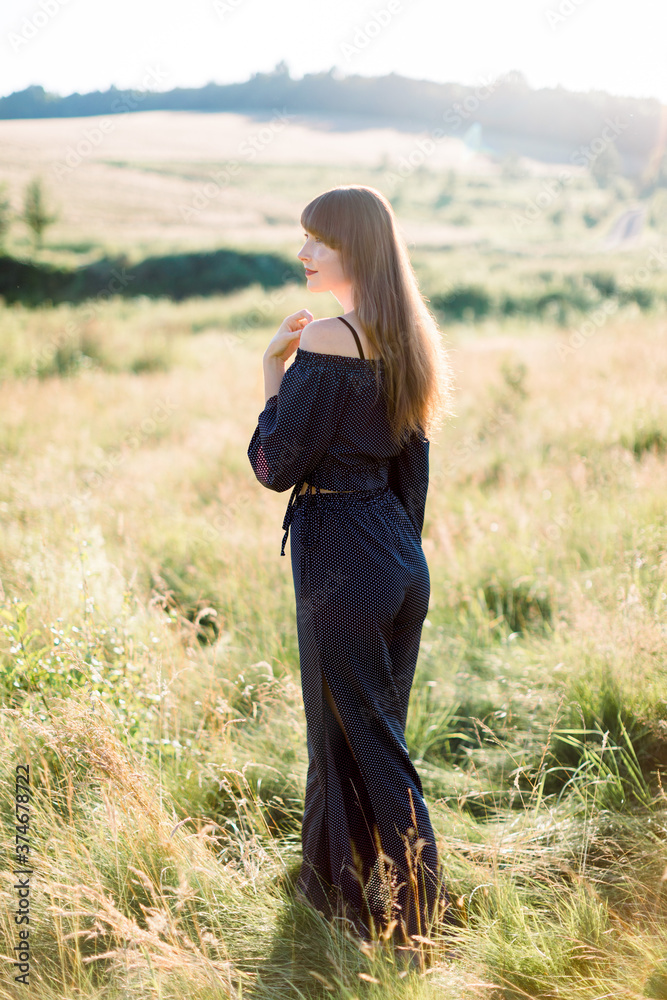 Happy young elegant dark haired woman in black clothes, enjoying nature, posing in beautiful summer green field at sunset. Pretty girl outdoor in the field