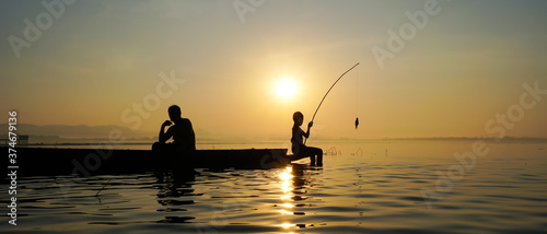 At lake side, asian fisherman sitting on boat while his son standing and using fishing rod to catch fish at the sunrise