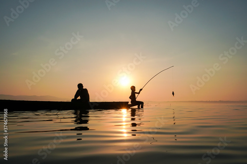 At lake side, asian fisherman sitting on boat while his son standing and  using fishing rod to catch fish at the sunrise Stock Photo